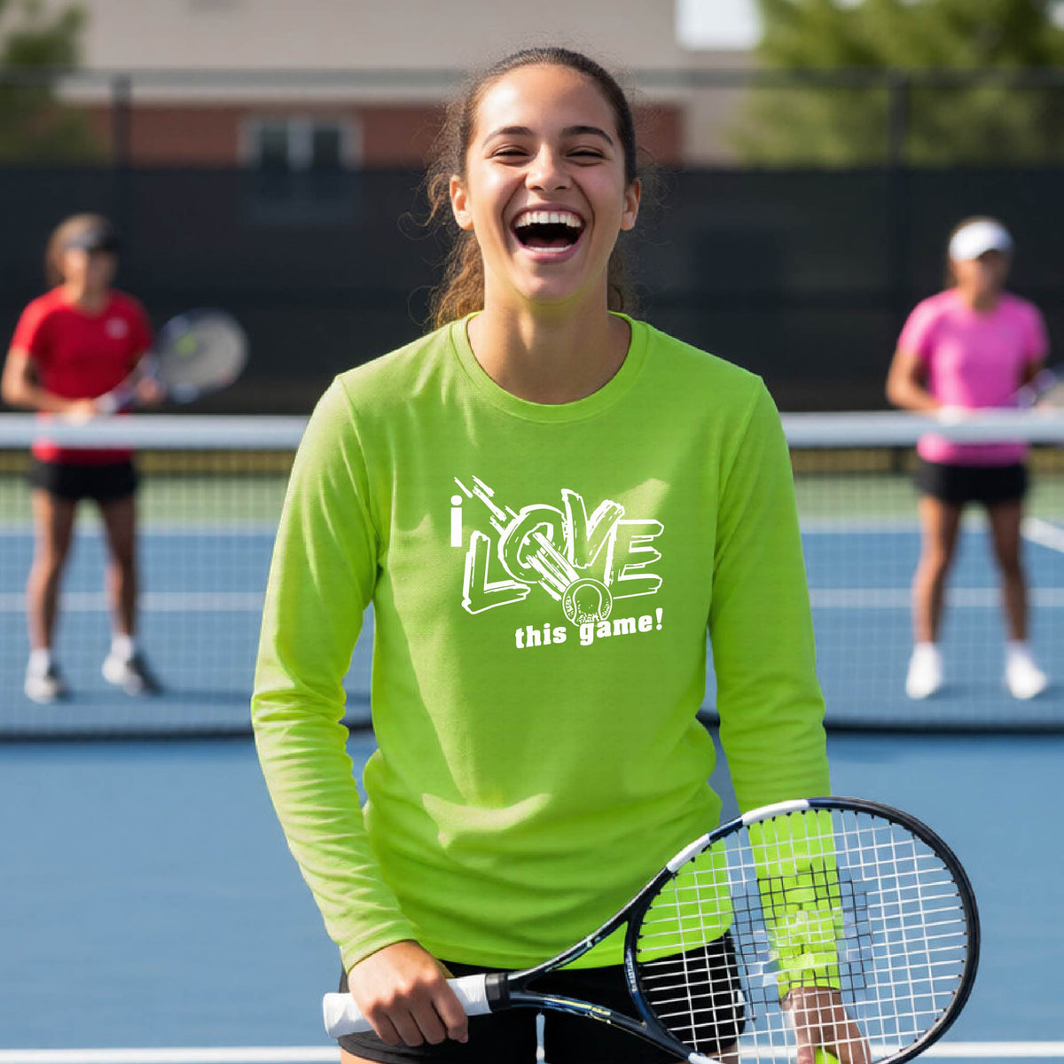 A woman laughing on a tennis court wearing a bright lime green long-sleeve shirt with the text 'I LOVE this game!' printed on the front, holding a tennis racket. Two other players are visible in the background.