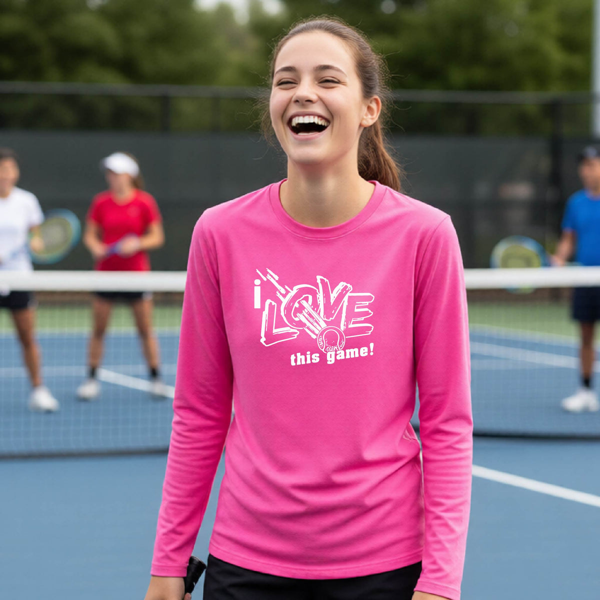 A woman laughing on a tennis court wearing a bright pink long-sleeve athletic shirt with a white graphic that reads 'I LOVE this game!'.
