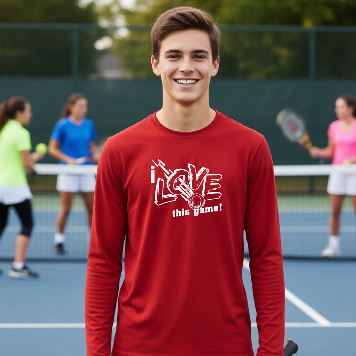 A smiling young man stands on a tennis court wearing a red long-sleeve shirt with a white graphic that reads 'I LOVE this game!'. Other tennis players are blurred in the background.