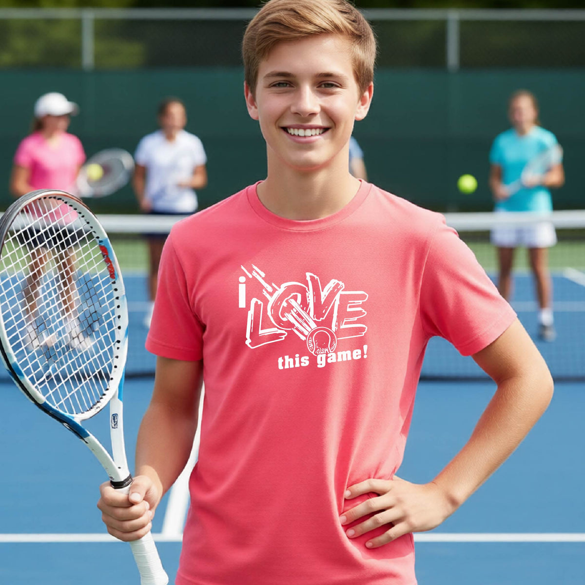 A smiling young man on a tennis court wearing a coral pink t-shirt with a white graphic that reads 'i LOVE this game!'. He is holding a tennis racket, with other players and a green tennis net visible in the background.