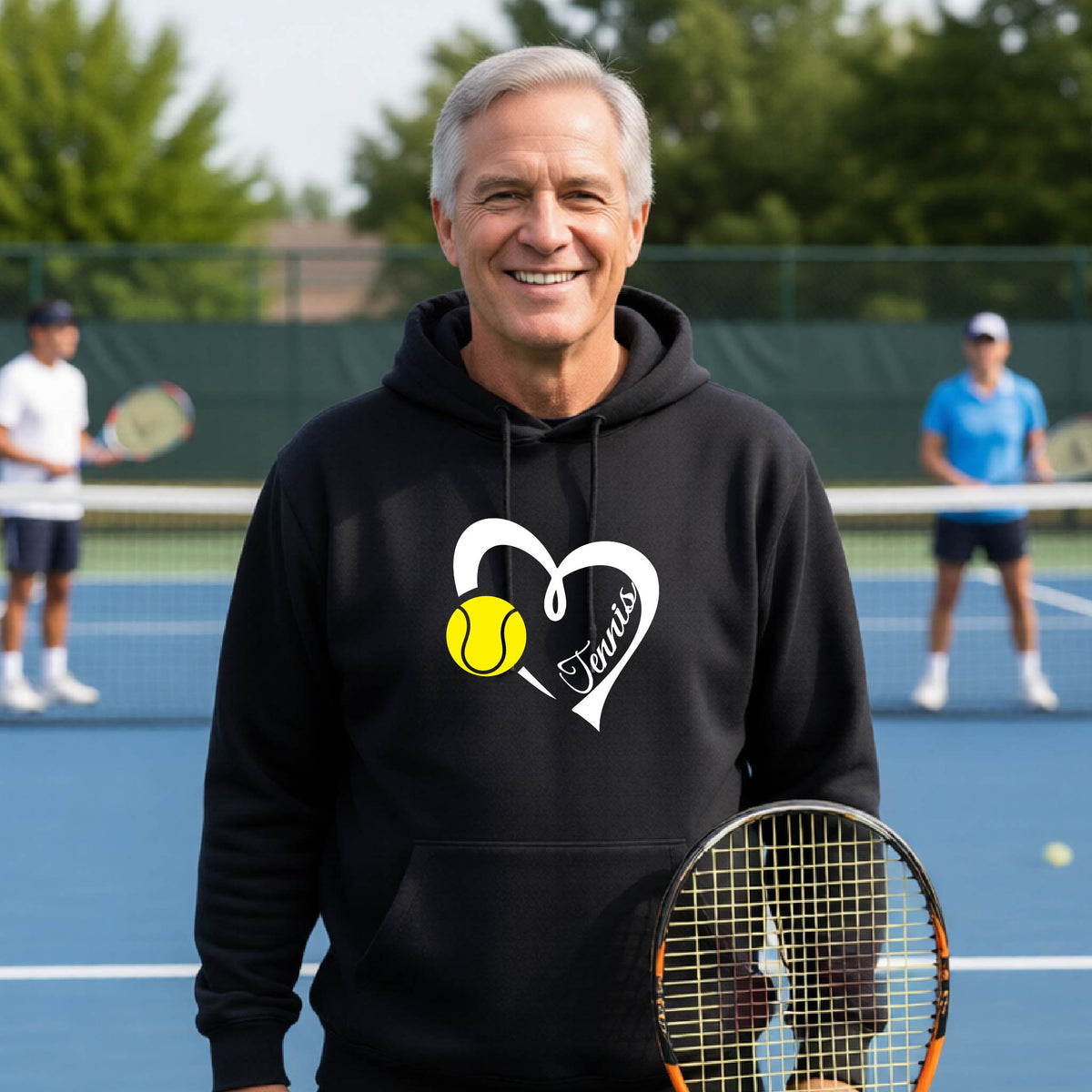 A smiling man wearing a black hoodie stands on a blue tennis court holding a racket. The hoodie has a white heart-shaped graphic with a yellow tennis ball and the word 'Tennis'.
