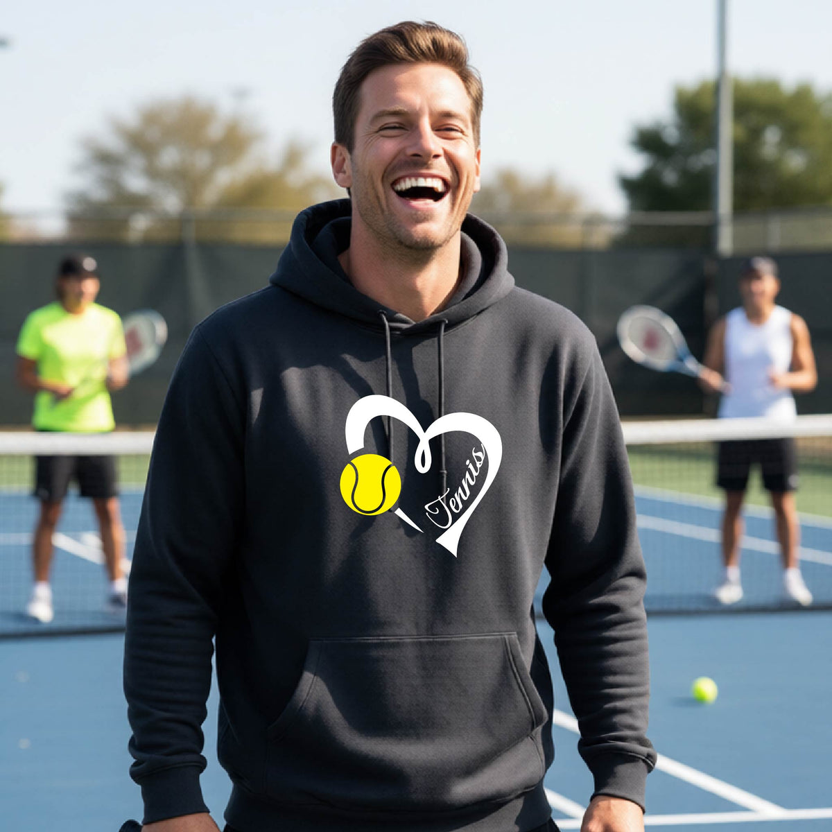 A man wearing a dark gray hoodie with a white heart graphic, a yellow tennis ball, and the name 'Tennis printed on the front, standing on a blue tennis court with two other players blurred in the background.