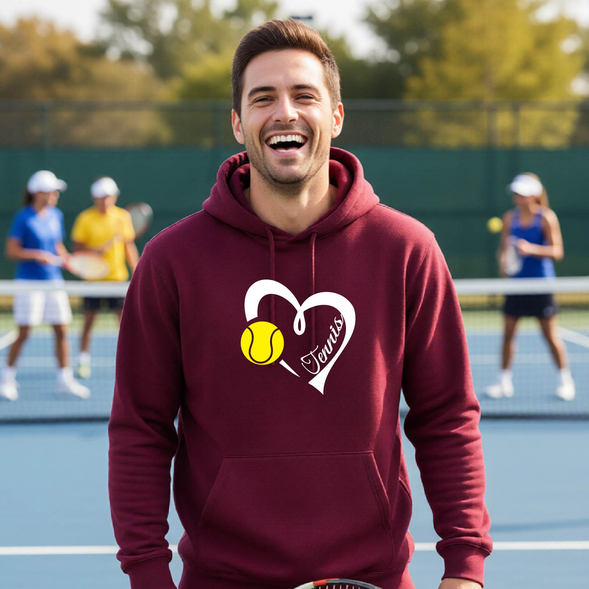 A man wearing a maroon hoodie with a white heart, yellow tennis ball, and the name 'Tennis' printed on the front, standing on a tennis court with other players blurred in the background.