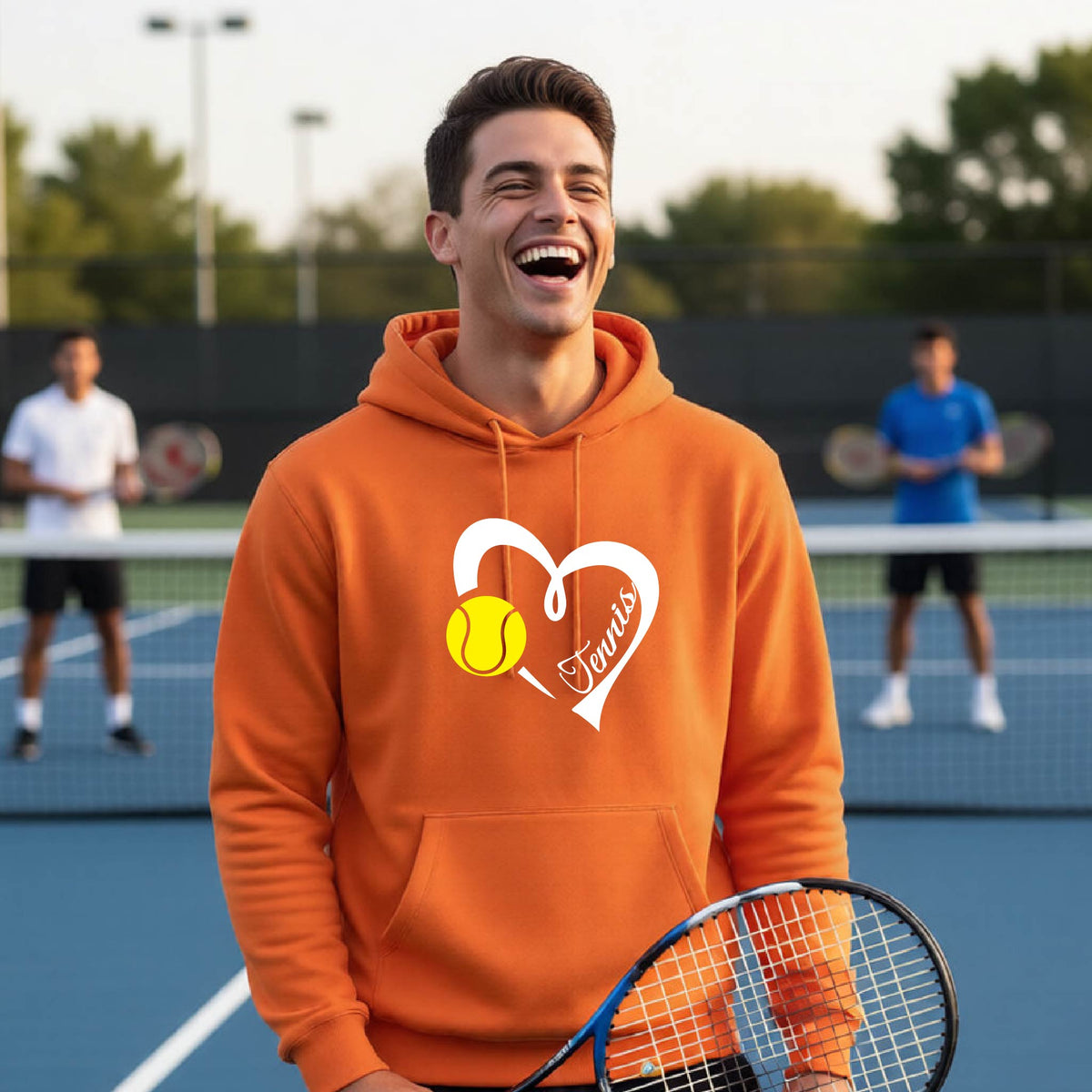 A man laughing on a tennis court wearing an orange hoodie with a white heart graphic, a yellow tennis ball, and the word 'Tennis' on the front.