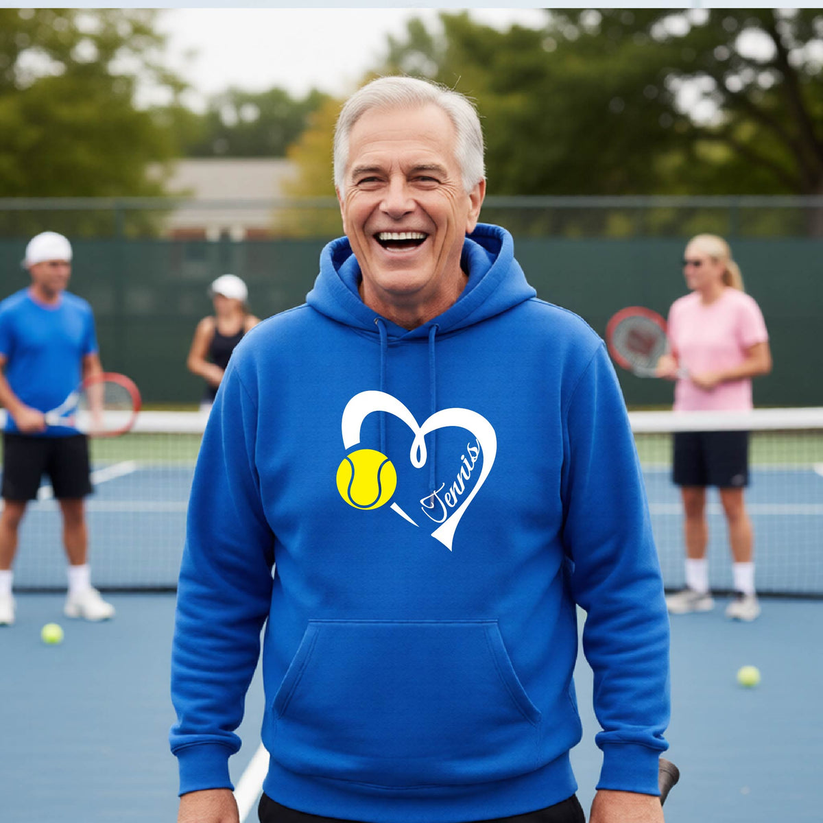 A smiling older man wearing a bright blue hoodie stands on a tennis court. The hoodie has a white heart outline with the word "Tennis" inside and a yellow tennis ball graphic over the chest.