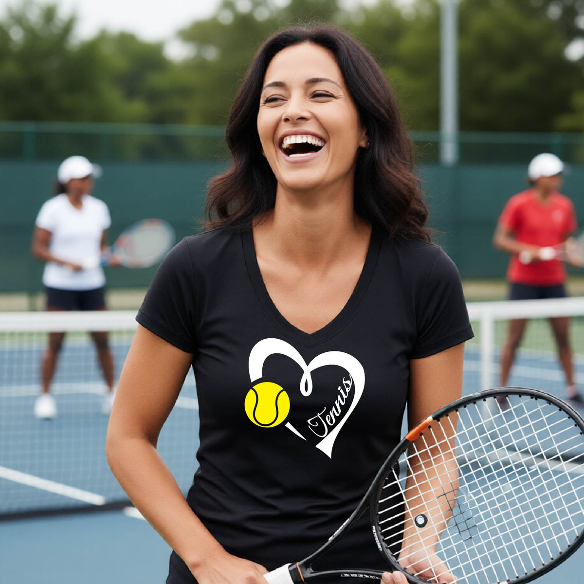 A woman laughing on a tennis court wearing a black V-neck t-shirt with a white heart outline, a yellow tennis ball graphic, and the word 'Tennis' inside. She is holding a tennis racket, with other players blurred in the background.
