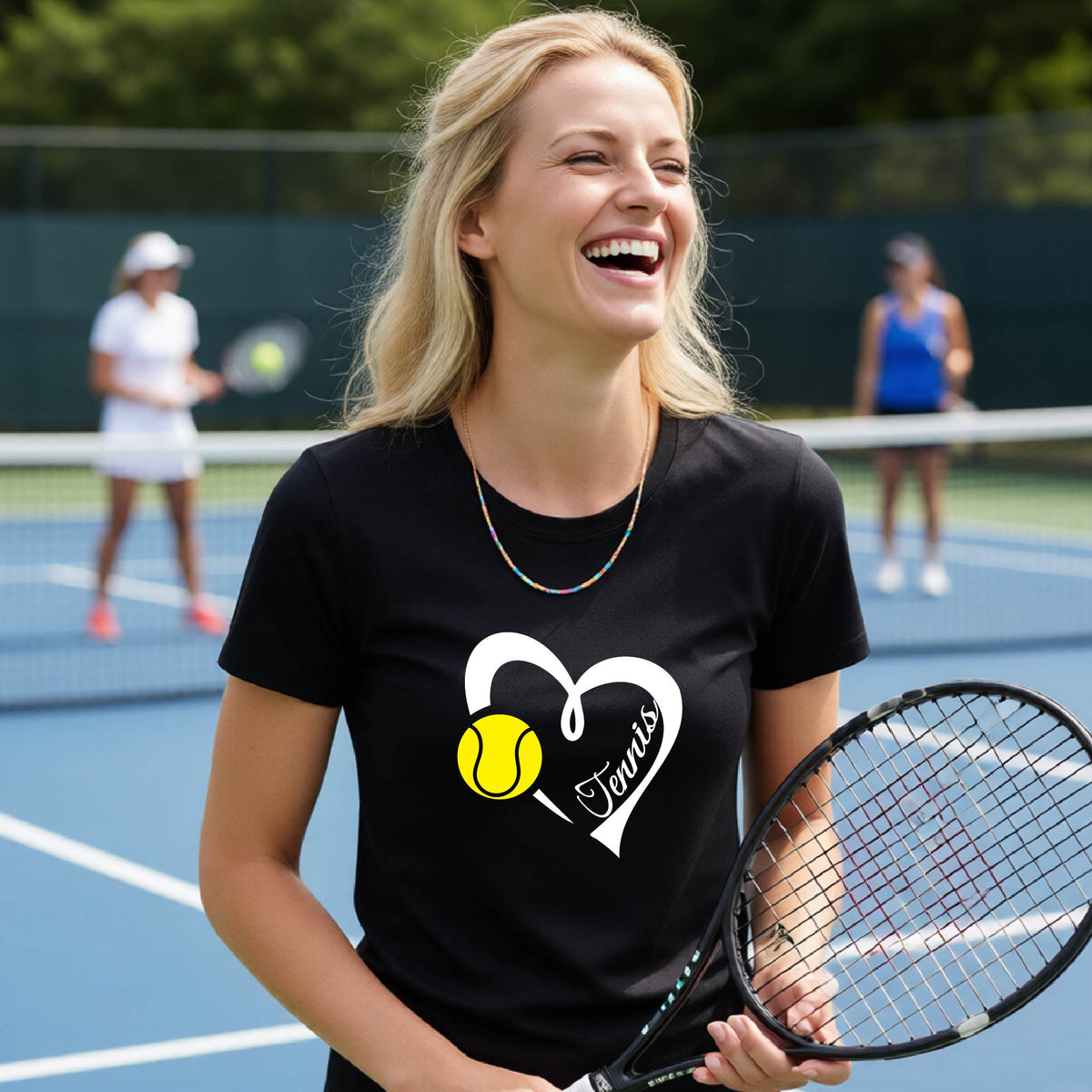 A woman on a blue tennis court wearing a black graphic t-shirt with a white heart, yellow tennis ball, and the word 'Tennis' in script. She is holding a tennis racket, with other players blurred in the background.