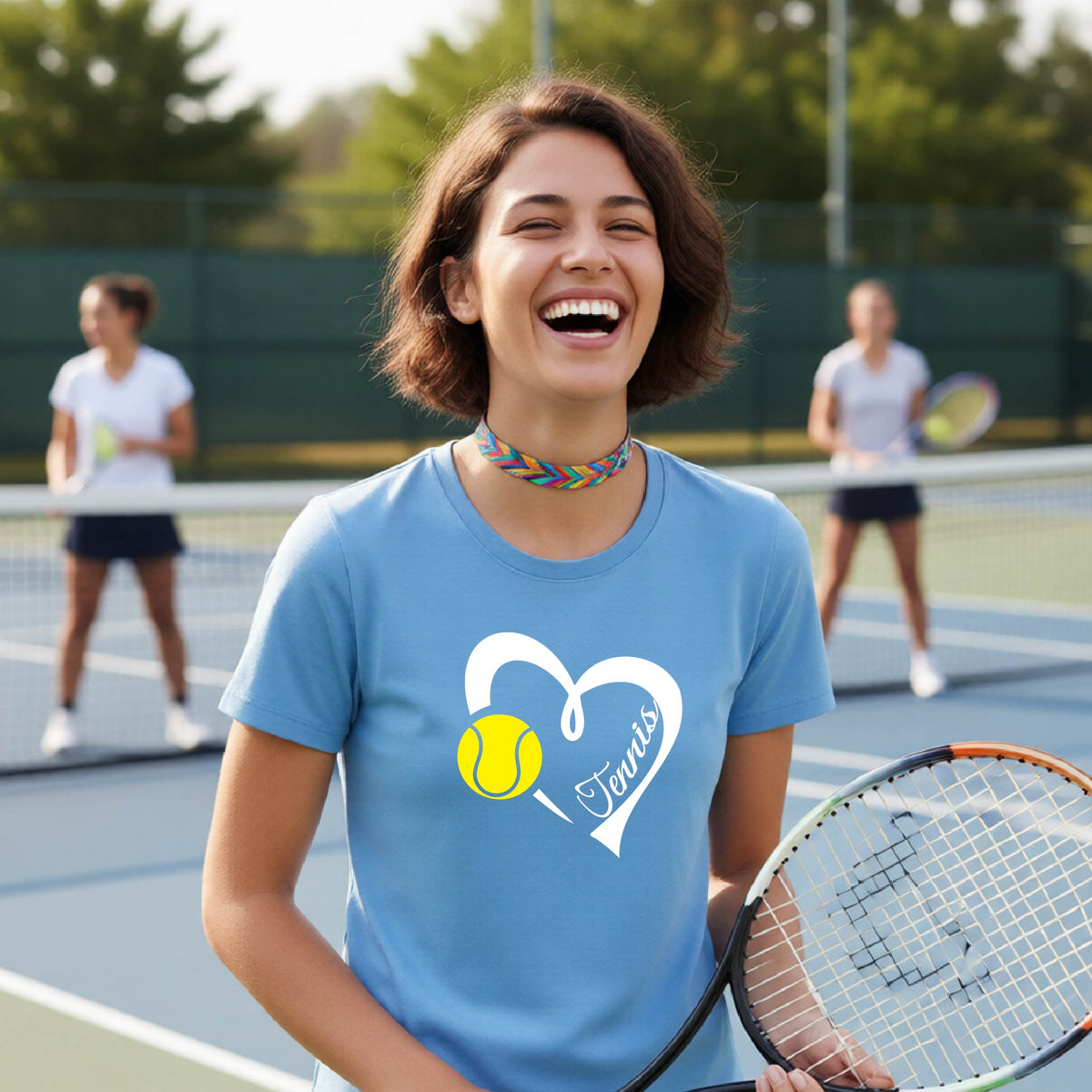 A woman laughing on a tennis court wearing a light blue t-shirt with a white heart graphic, a yellow tennis ball, and the word 'Tennis' written inside. Two other players are blurred in the background.