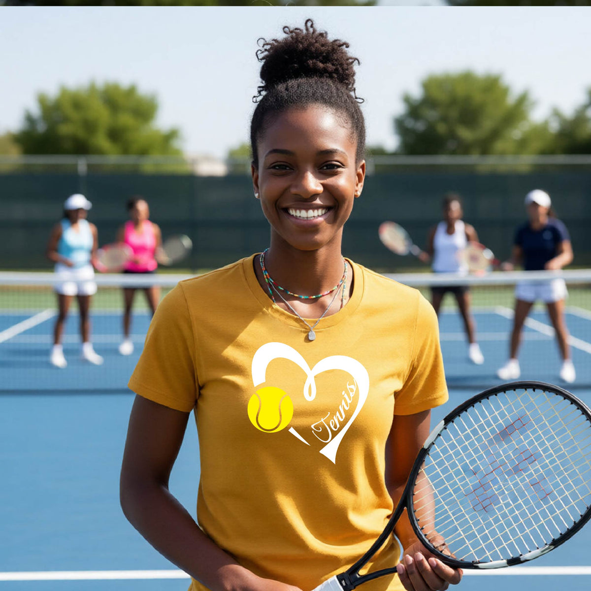 A smiling woman stands on a blue tennis court holding a tennis racket, wearing a mustard yellow graphic T-shirt with a white heart, tennis ball, and the word 'Tennis'. Other players are blurred in the background.