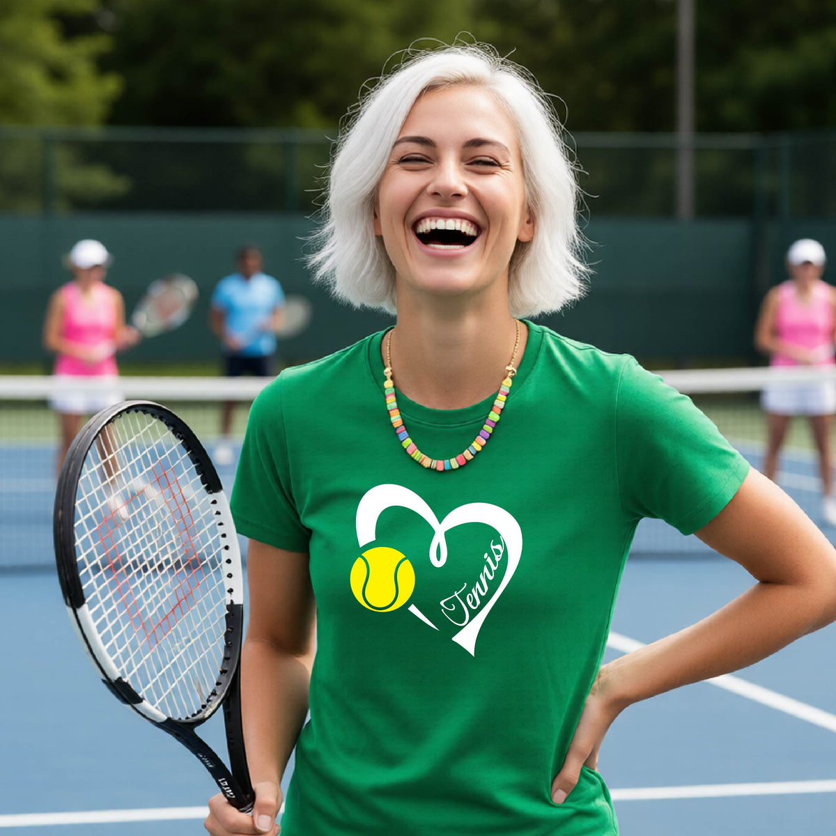 A woman with short white hair laughing on a tennis court, wearing a green t-shirt with a white heart outline, a tennis ball graphic, and the word 'Tennis'. She is holding a tennis racket, with other players blurred in the background.