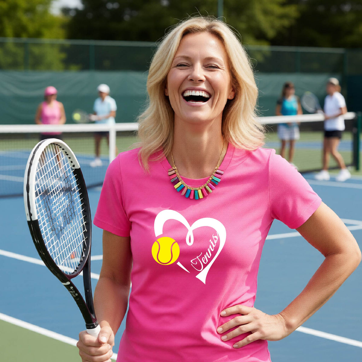 A smiling blonde woman on a tennis court wearing a bright pink t-shirt with a white heart graphic, a yellow tennis ball, and the name 'Tennis' printed on the front. She is holding a tennis racket, with other players and the court visible in the background.