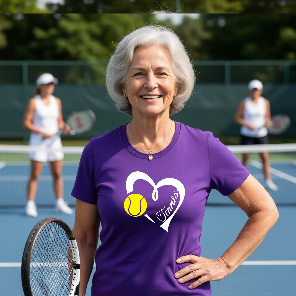 An older woman with gray hair stands on a tennis court wearing a purple short-sleeve T-shirt with a white heart outline, a yellow tennis ball graphic, and the word 'Tennis' inside. Two blurred tennis players are visible in the background.