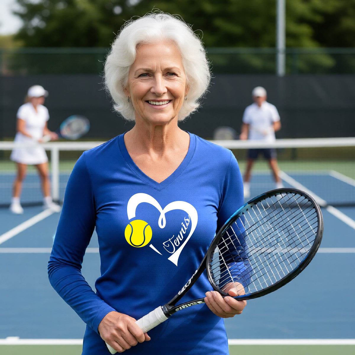 An older woman with white hair stands on a tennis court holding a tennis racket, wearing a blue long-sleeve V-neck shirt with a white heart outline, a yellow tennis ball graphic, and the word 'Tennis' printed on the front. Two other players are blurred in the background.