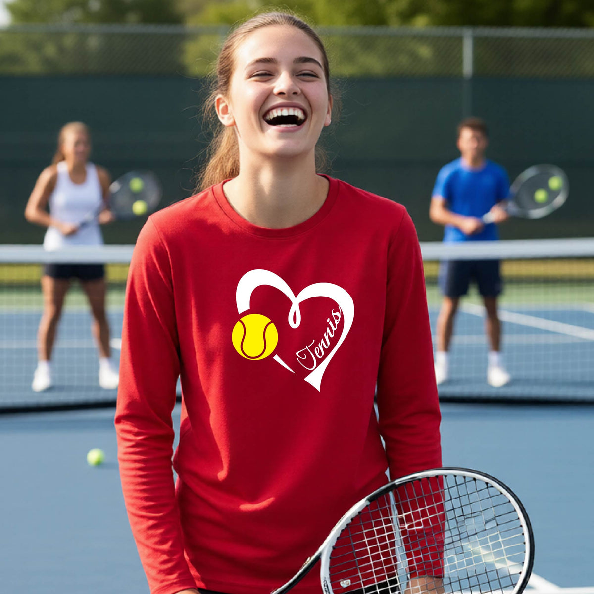 A woman on a tennis court wearing a red long-sleeve shirt with a white heart outline, a yellow tennis ball graphic, and the word 'Tennis' in script. Two other tennis players are blurred in the background.