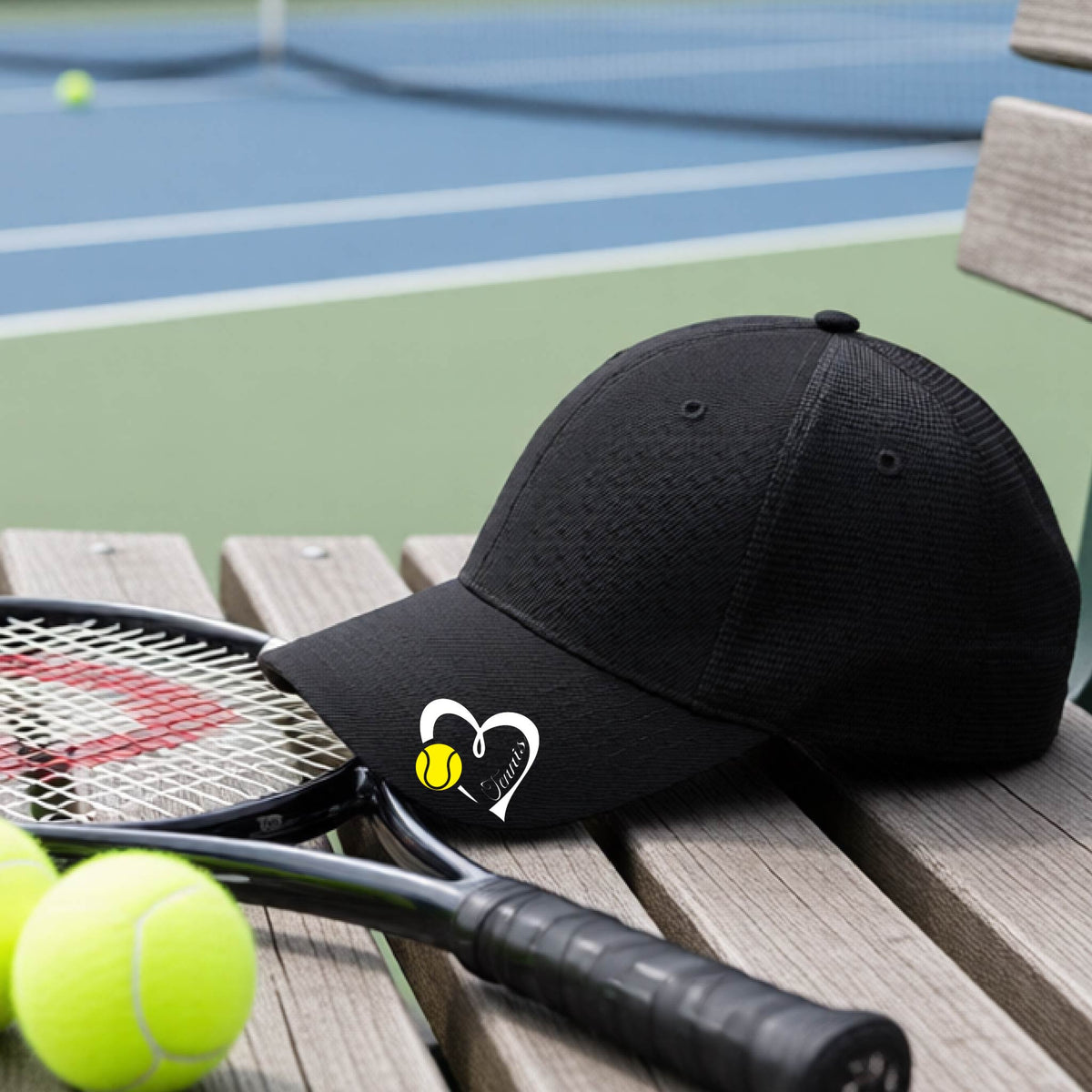 A black baseball cap with a white heart and tennis ball logo with 'Tennis' written in script inside the heart, on the brim of the hat, resting on a wooden bench at a tennis court with tennis balls and a racket nearby.