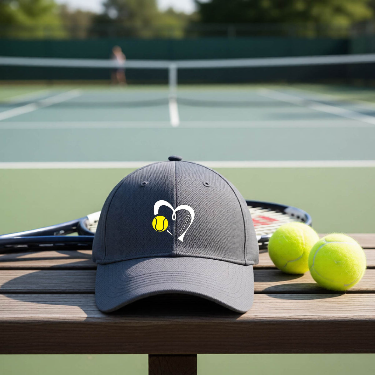 A graphite gray baseball cap with a white heart and tennis ball logo with 'Tennis' written in script inside the heart, on the front of the hat, resting on a wooden bench at a tennis court with tennis balls and a racket nearby.
