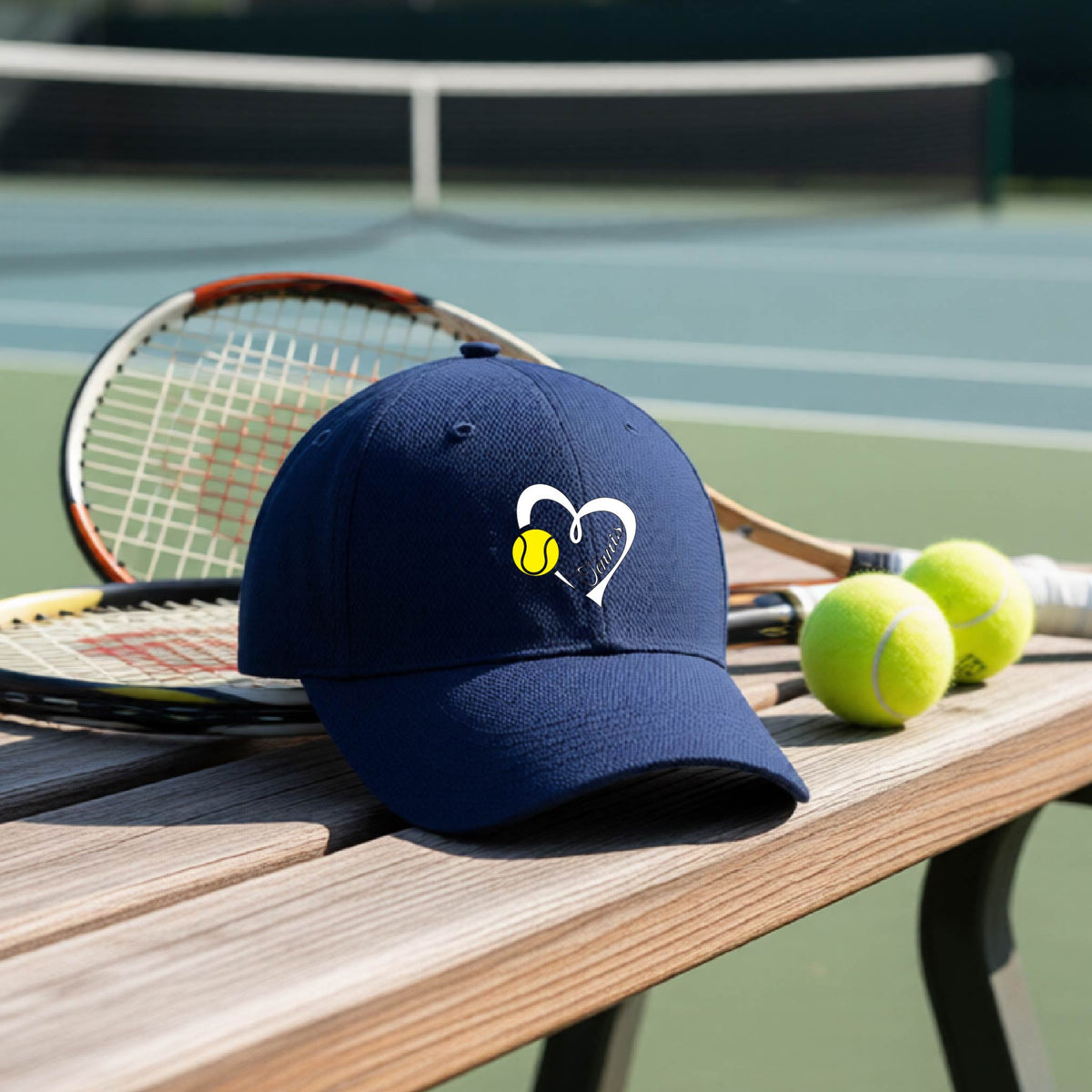 A navy blue baseball cap with a white heart and tennis ball logo with 'Tennis' written in script inside the heart, on the front of the hat, resting on a wooden bench at a tennis court with tennis balls and a racket nearby.