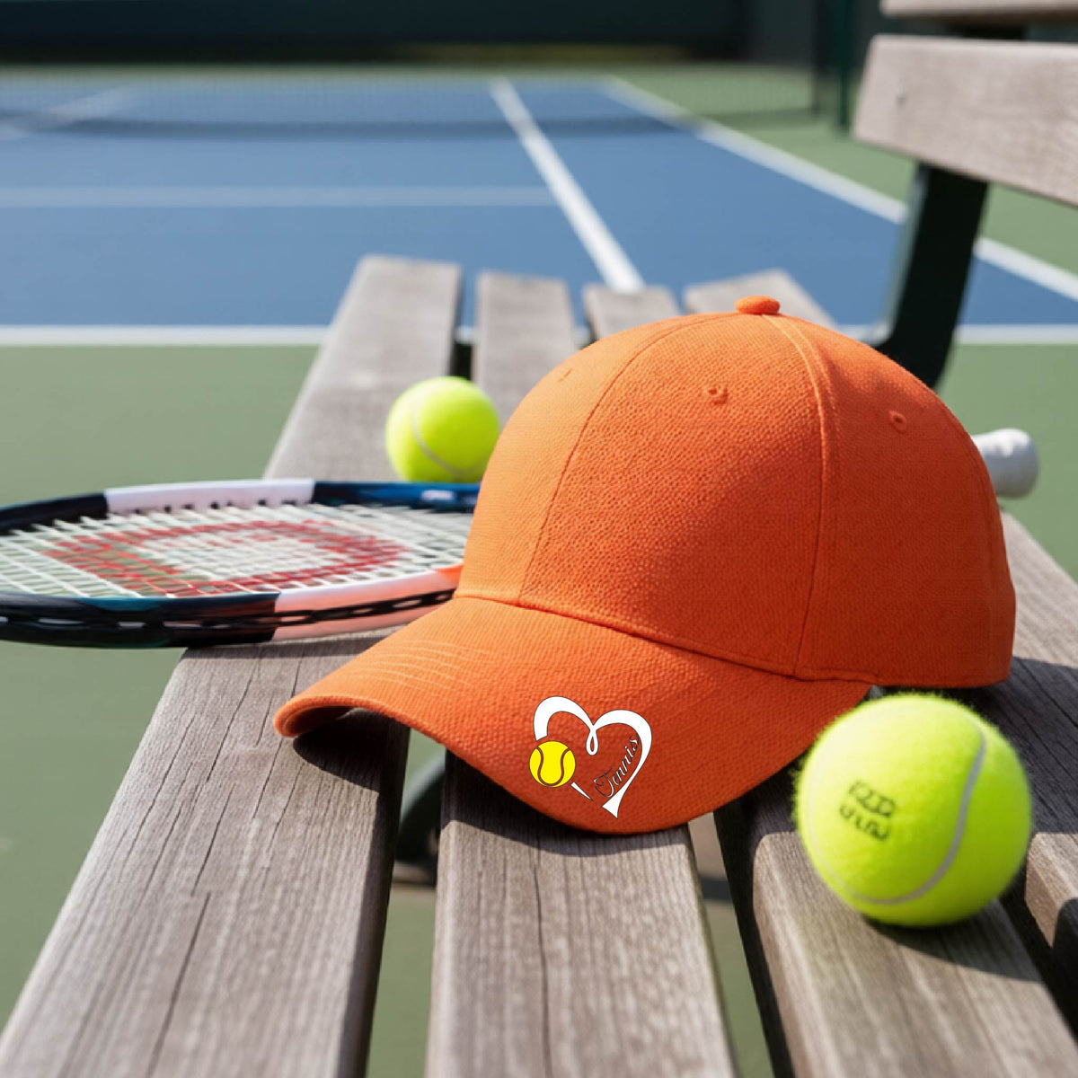 An orange baseball cap with a white heart and tennis ball logo with 'Tennis' written in script inside the heart, on the brim of the hat, resting on a wooden bench at a tennis court with tennis balls and a racket nearby.