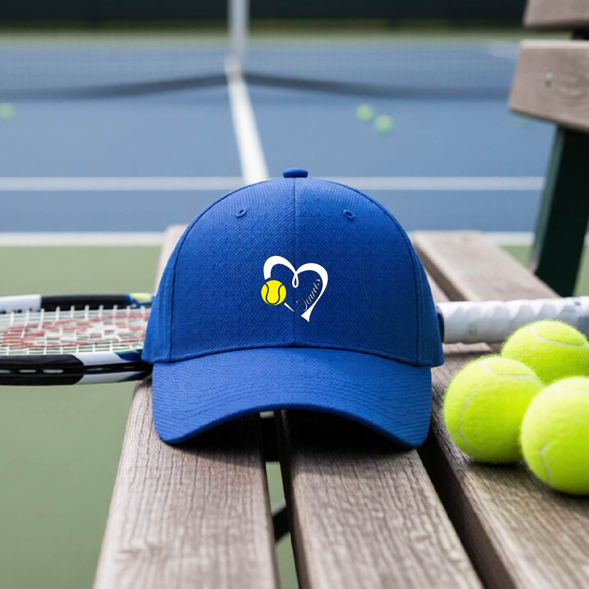 A royal blue baseball cap with a white heart and tennis ball logo with 'Tennis' written in script inside the heart, on the front of the hat, resting on a wooden bench at a tennis court with tennis balls and a racket nearby.