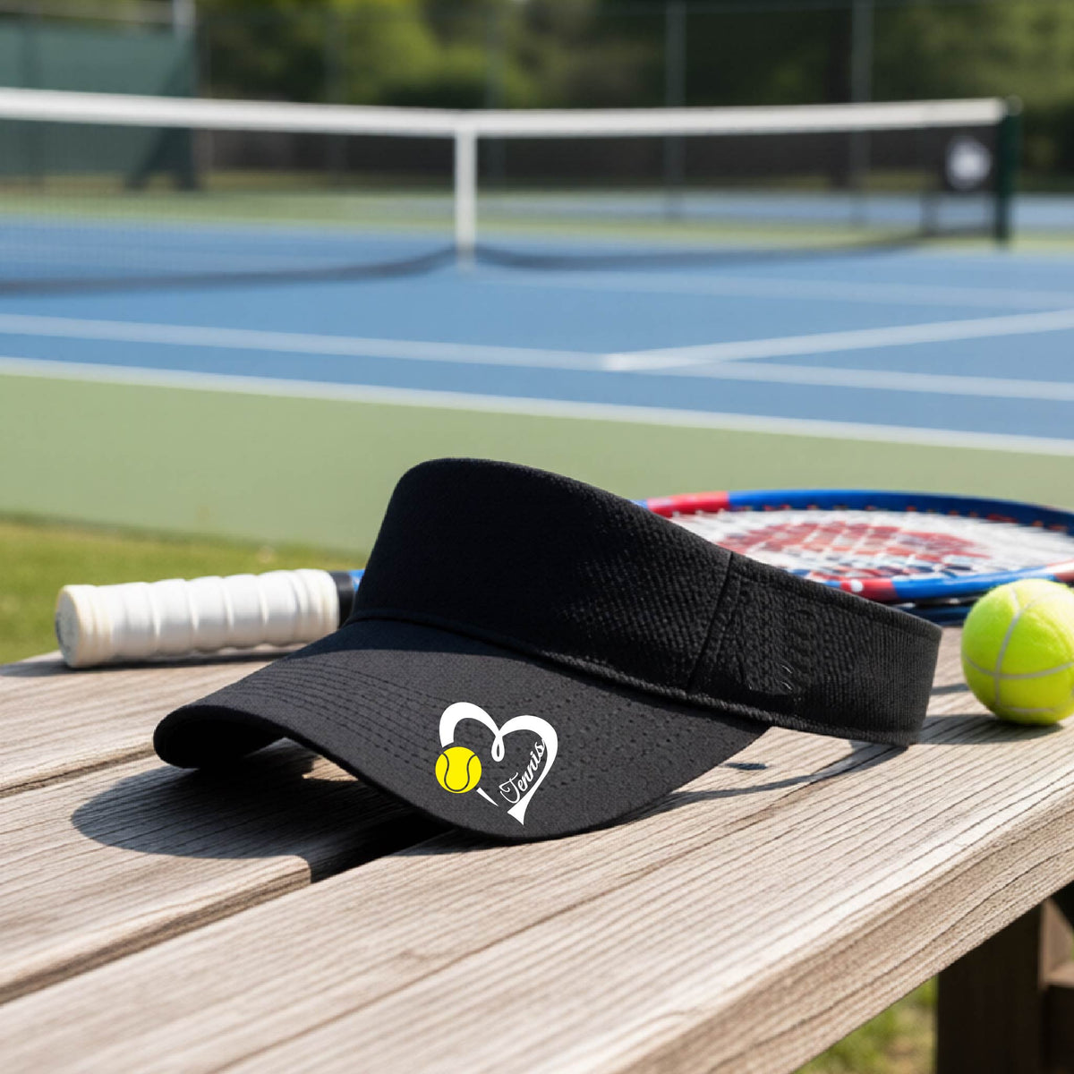 A black visor with a white heart and tennis ball graphic with the 'Tennis' written in white script inside the hearts, on the brim of the visor, sits on a wooden bench at a tennis court, with tennis rackets and yellow balls nearby.