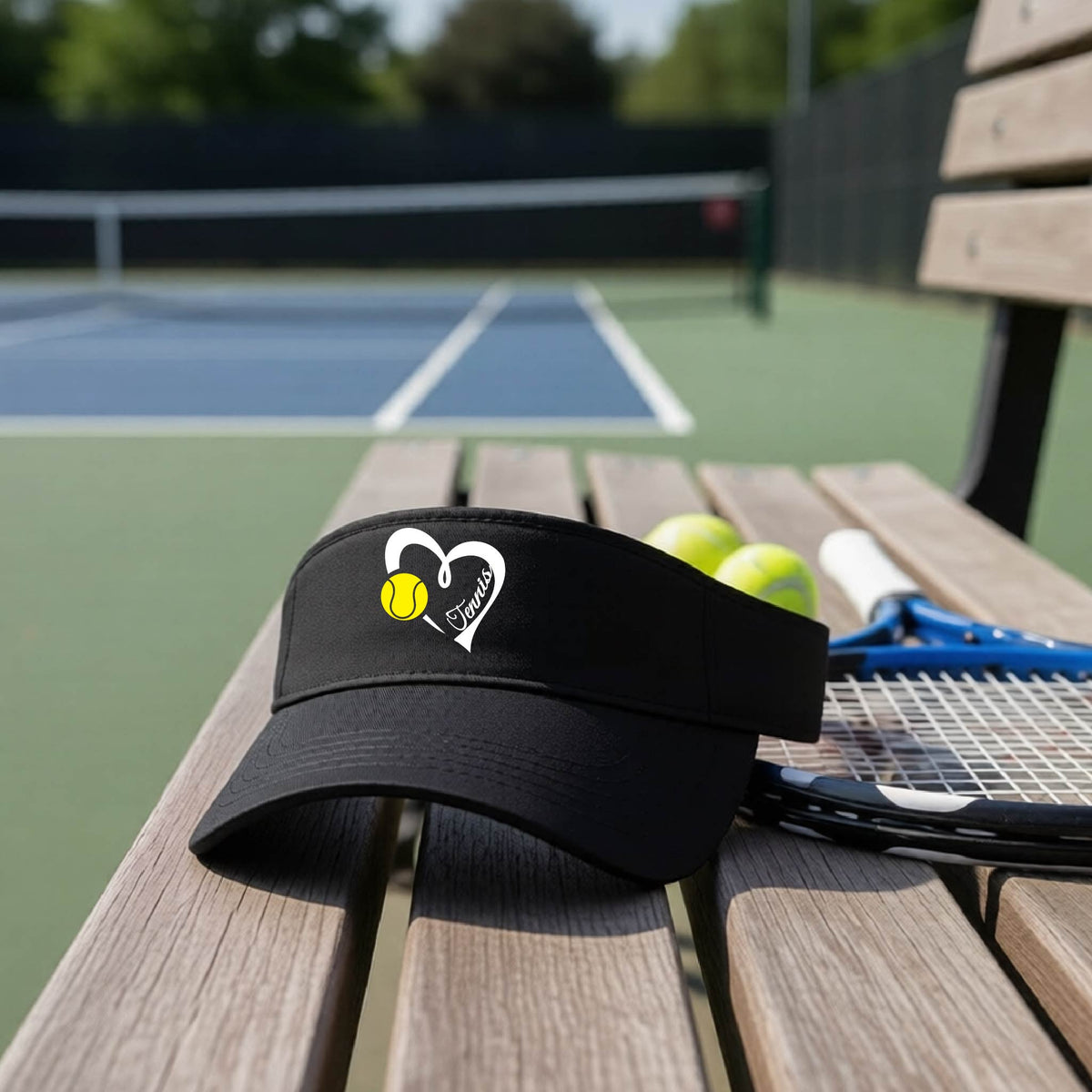 A black visor with a white heart and tennis ball graphic with the 'Tennis' written in white script inside the hearts, on the front of the visor, sits on a wooden bench at a tennis court, with tennis rackets and yellow balls nearby.