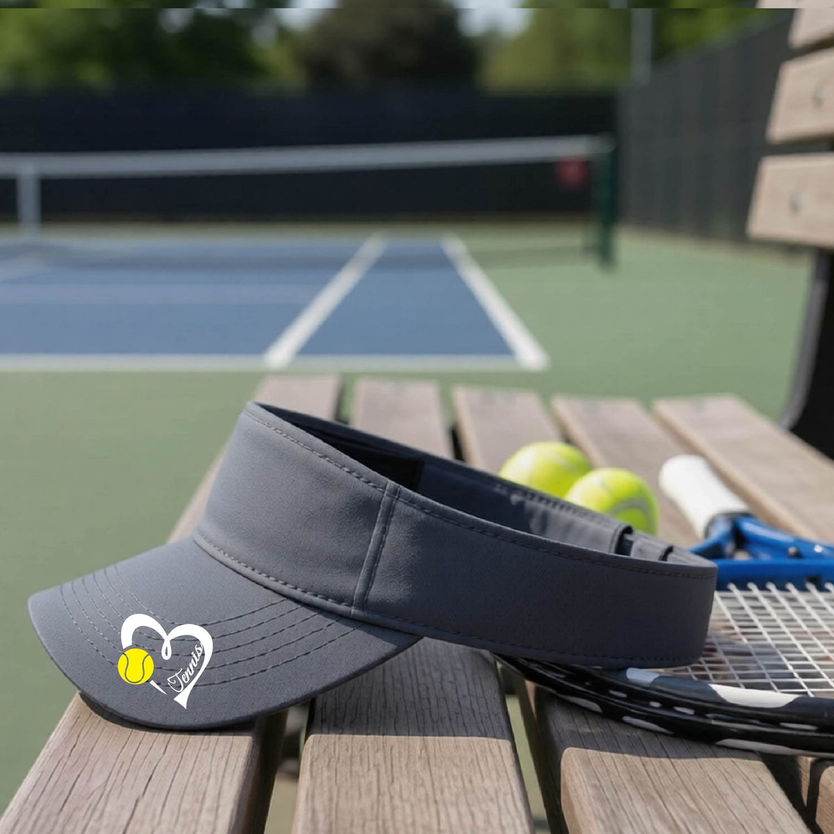 A dark gray visor with a white heart and tennis ball graphic with the 'Tennis' written in white script inside the hearts, on the brim of the visor, sits on a wooden bench at a tennis court, with tennis rackets and yellow balls nearby.