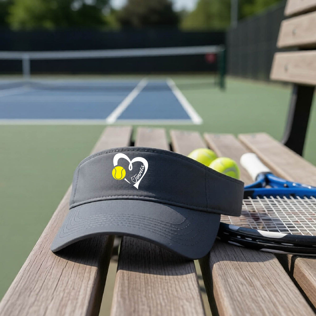 A dark gray visor with a white heart and tennis ball graphic with the 'Tennis' written in white script inside the hearts, on the front of the visor, sits on a wooden bench at a tennis court, with tennis rackets and yellow balls nearby.