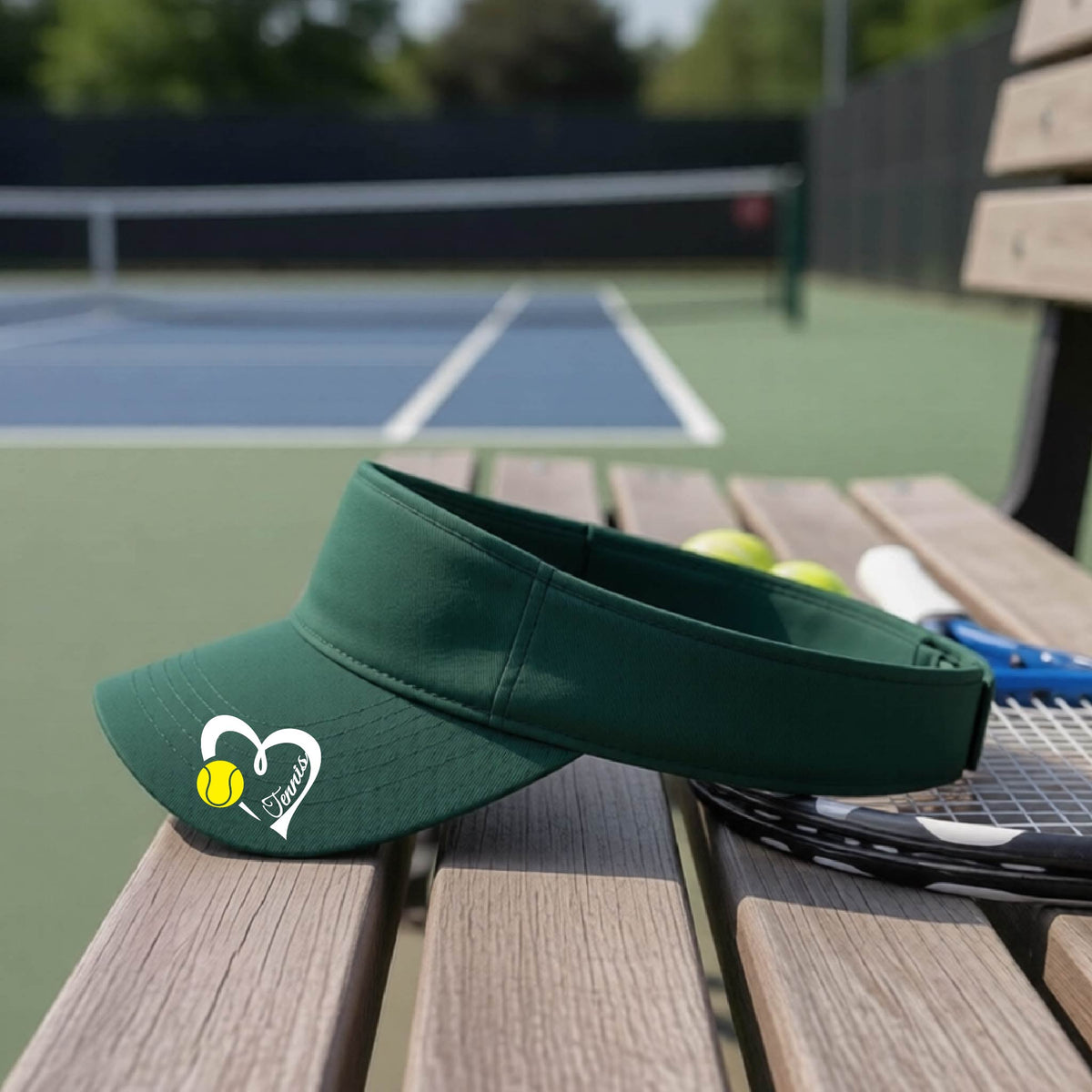 A dark green visor with a white heart and tennis ball graphic with the 'Tennis' written in white script inside the hearts, on the brim of the visor, sits on a wooden bench at a tennis court, with tennis rackets and yellow balls nearby.