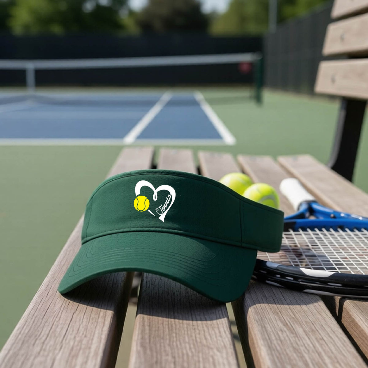A dark green visor with a white heart and tennis ball graphic with the 'Tennis' written in white script inside the hearts, on the front of the visor, sits on a wooden bench at a tennis court, with tennis rackets and yellow balls nearby.