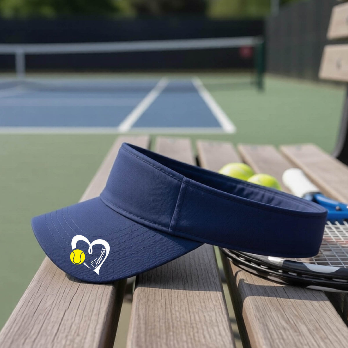 A navy blue visor with a white heart and tennis ball graphic with the 'Tennis' written in white script inside the hearts, on the brim of the visor, sits on a wooden bench at a tennis court, with tennis rackets and yellow balls nearby.