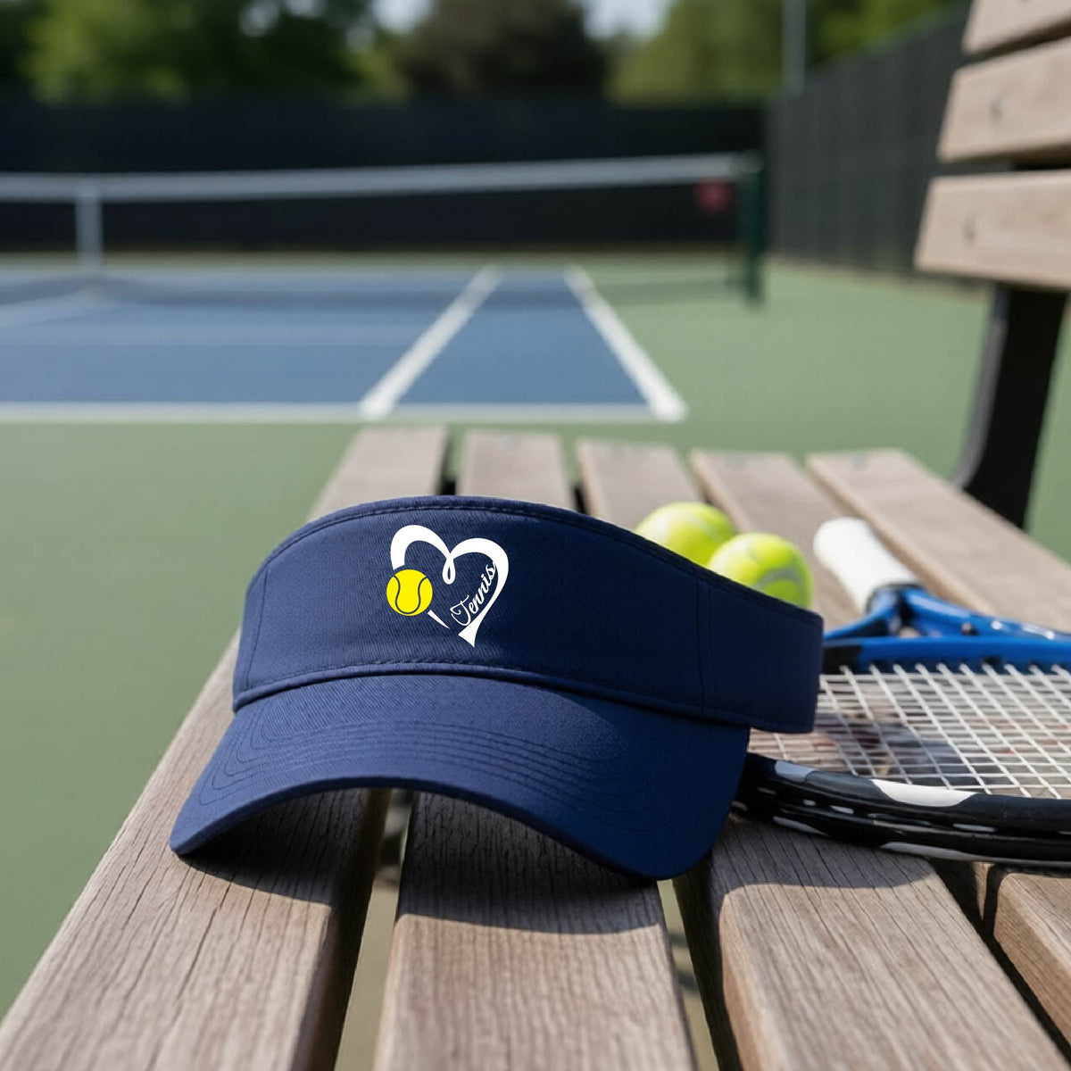 A navy blue visor with a white heart and tennis ball graphic with the 'Tennis' written in white script inside the hearts, on the front of the visor, sits on a wooden bench at a tennis court, with tennis rackets and yellow balls nearby.