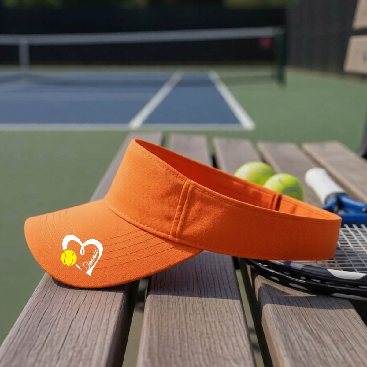 An orange visor with a white heart and tennis ball graphic with the 'Tennis' written in white script inside the hearts, on the brim of the visor, sits on a wooden bench at a tennis court, with tennis rackets and yellow balls nearby.