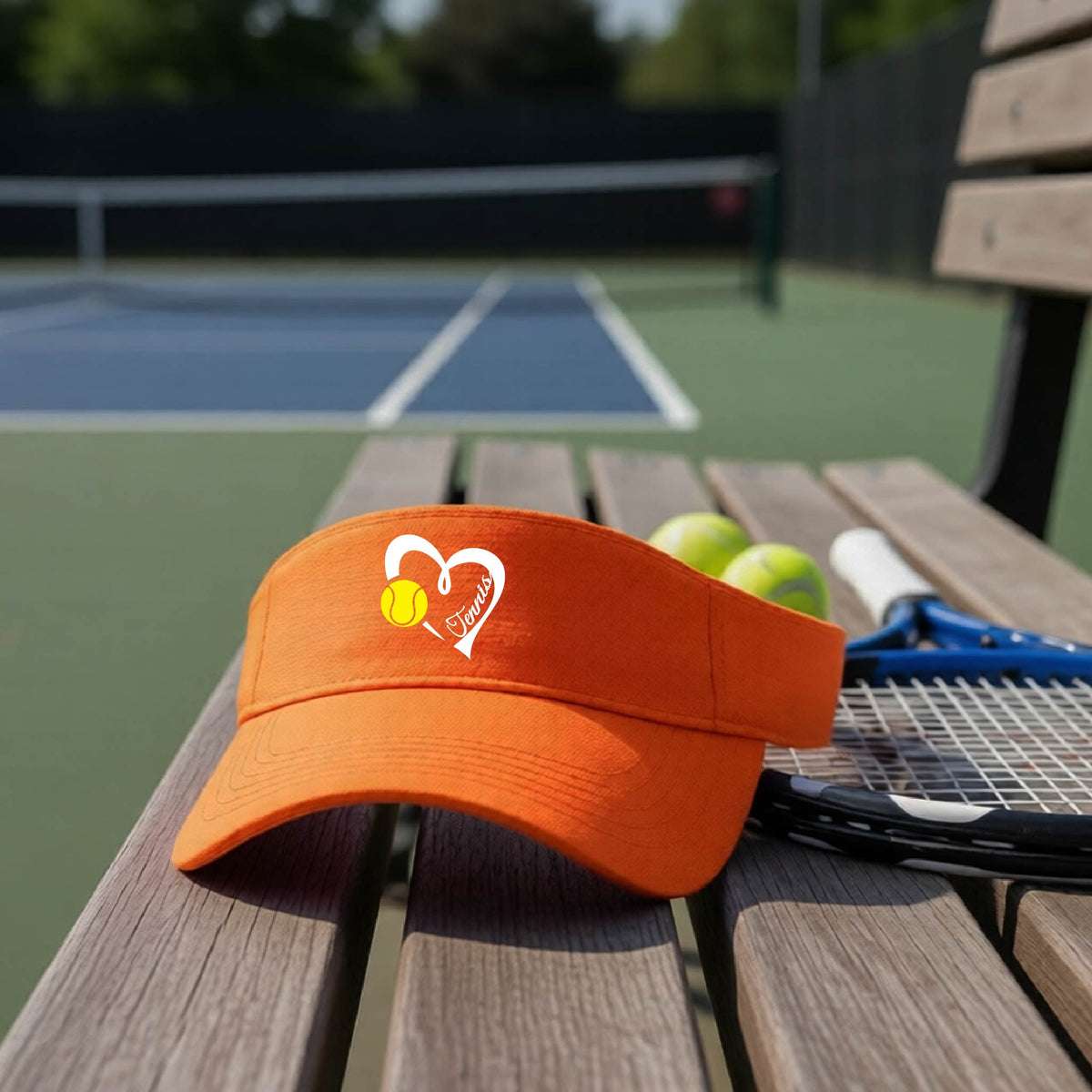 An orange visor with a white heart and tennis ball graphic with the 'Tennis' written in white script inside the hearts, on the front of the visor, sits on a wooden bench at a tennis court, with tennis rackets and yellow balls nearby.