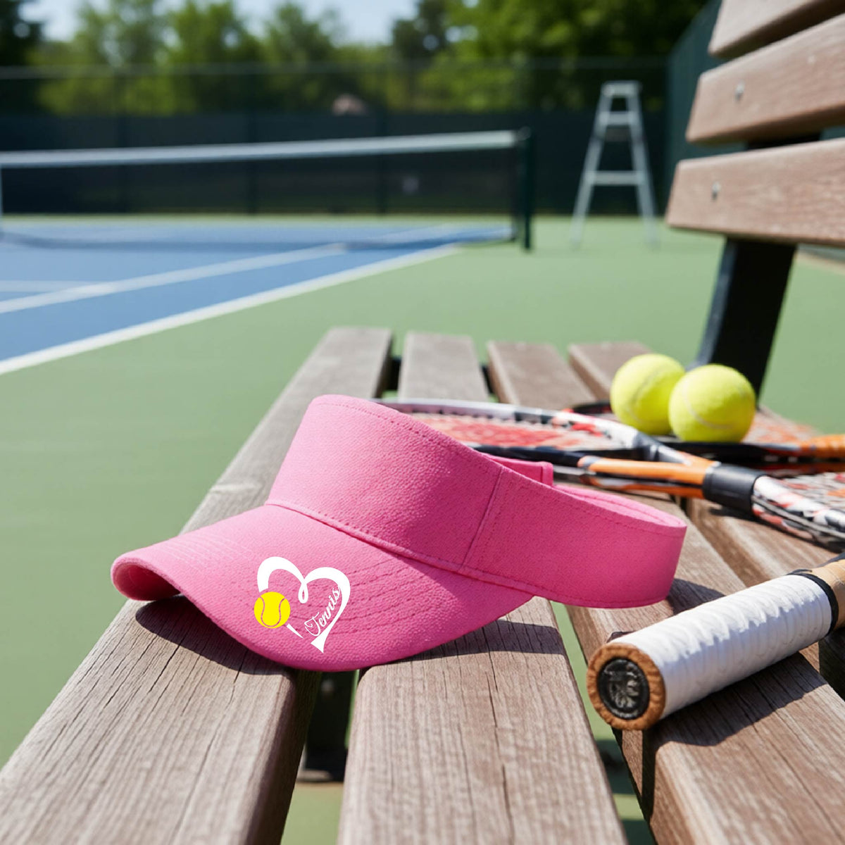 A pink visor with a white heart and tennis ball graphic with the 'Tennis' written in white script inside the hearts, on the brim of the visor, sits on a wooden bench at a tennis court, with tennis rackets and yellow balls nearby.