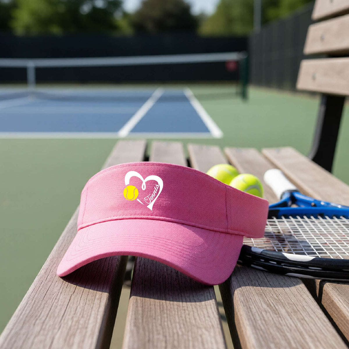 A pink visor with a white heart and tennis ball graphic with the 'Tennis' written in white script inside the hearts, on the front of the visor, sits on a wooden bench at a tennis court, with tennis rackets and yellow balls nearby.