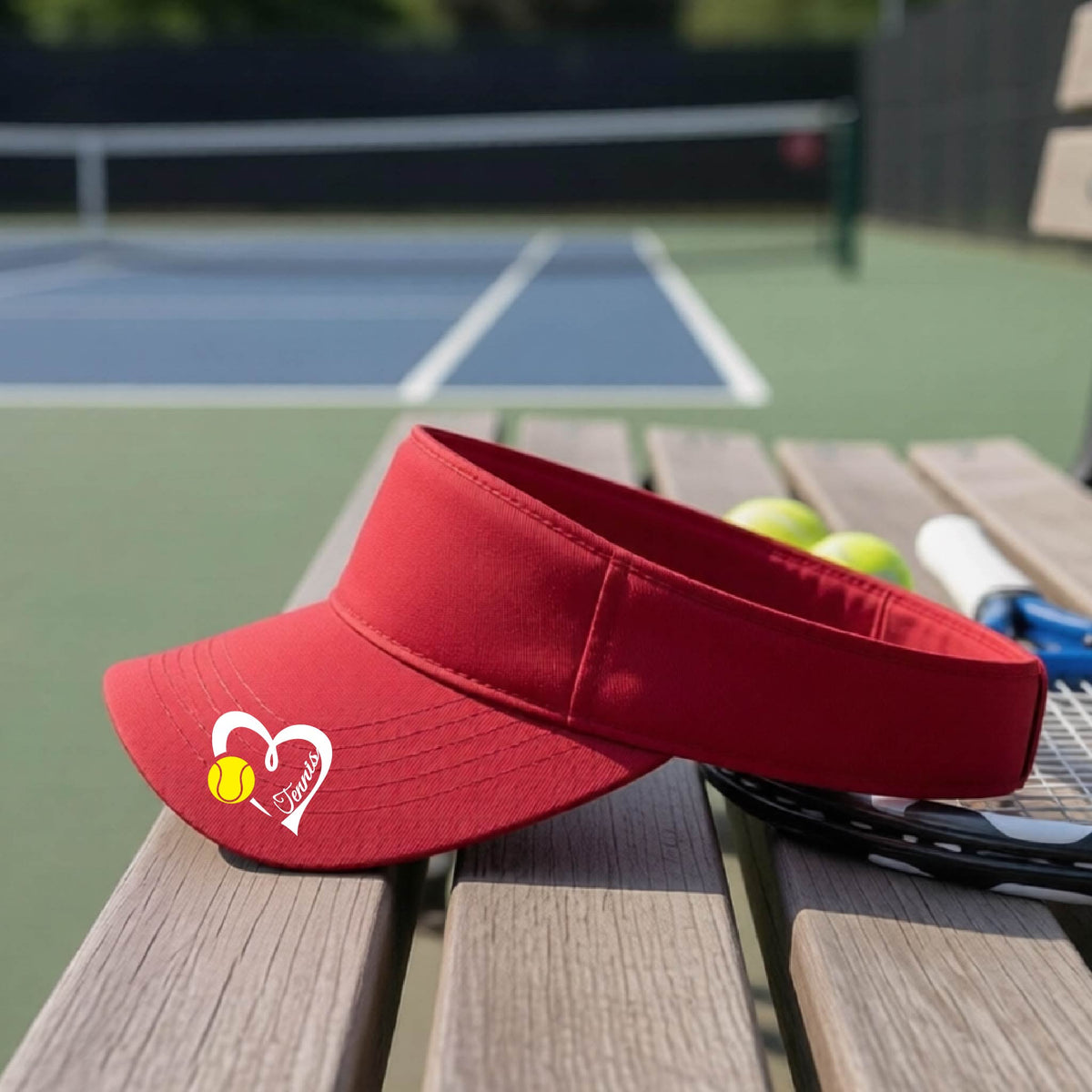 A red visor with a white heart and tennis ball graphic with the 'Tennis' written in white script inside the hearts, on the brim of the visor, sits on a wooden bench at a tennis court, with tennis rackets and yellow balls nearby.