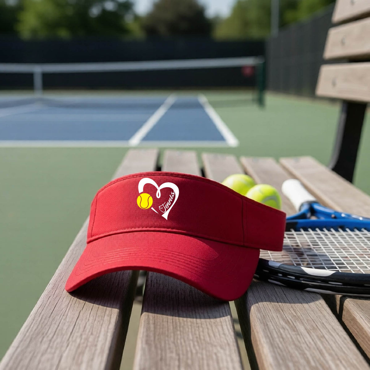 A red visor with a white heart and tennis ball graphic with the 'Tennis' written in white script inside the hearts, on the front of the visor, sits on a wooden bench at a tennis court, with tennis rackets and yellow balls nearby.