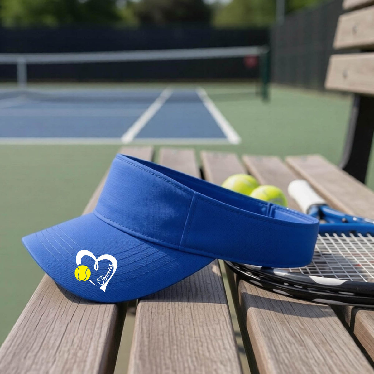 A royal blue visor with a white heart and tennis ball graphic with the 'Tennis' written in white script inside the hearts, on the brim of the visor, sits on a wooden bench at a tennis court, with tennis rackets and yellow balls nearby.