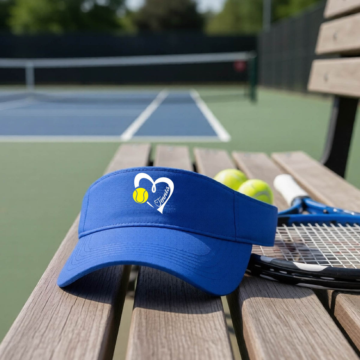 A royal blue visor with a white heart and tennis ball graphic with the 'Tennis' written in white script inside the hearts, on the front of the visor, sits on a wooden bench at a tennis court, with tennis rackets and yellow balls nearby.