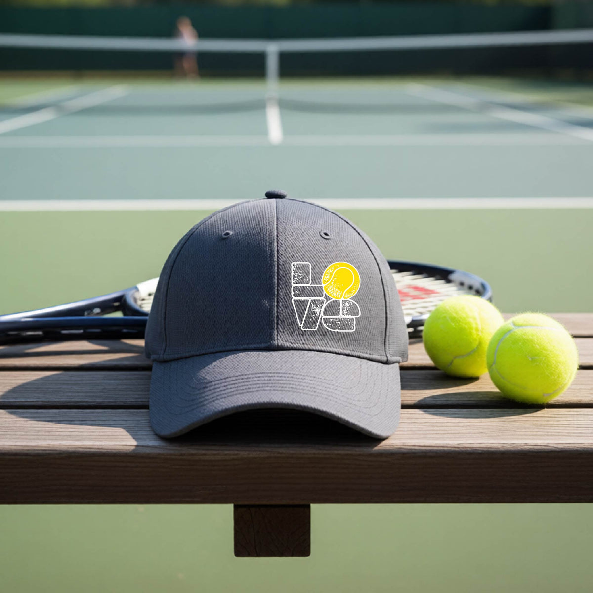 A graphite gray hat with a white and yellow tennis-themed graphic "LOVE" with a tennis ball replacing the O, sits on a wooden bench at a tennis court, with tennis balls and a racket in the background.