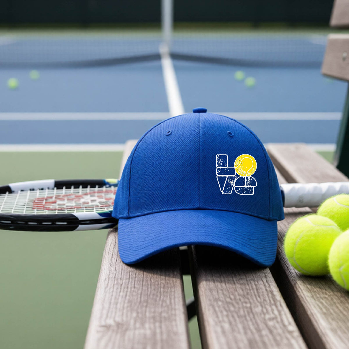 A royal blue hat with a white and yellow tennis-themed graphic "LOVE" with a tennis ball replacing the O, sits on a wooden bench at a tennis court, with tennis balls and a racket in the background.