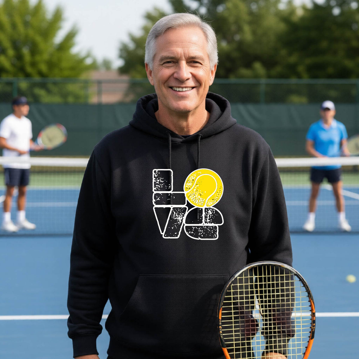 A smiling man wearing a black hoodie with a 'LOVE' tennis graphic with a tennis ball replacing the O, stands on an outdoor tennis court, with other players blurred in the background