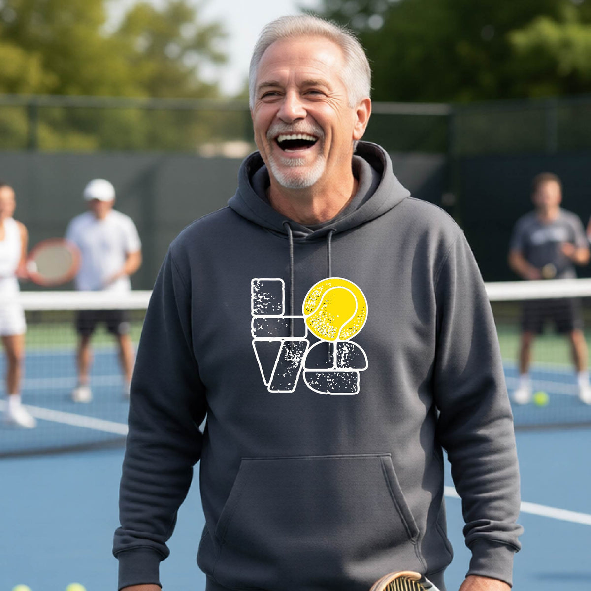 A smiling man wearing a dark gray hoodie with a 'LOVE' tennis graphic with a tennis ball replacing the O, stands on an outdoor tennis court, with other players blurred in the background