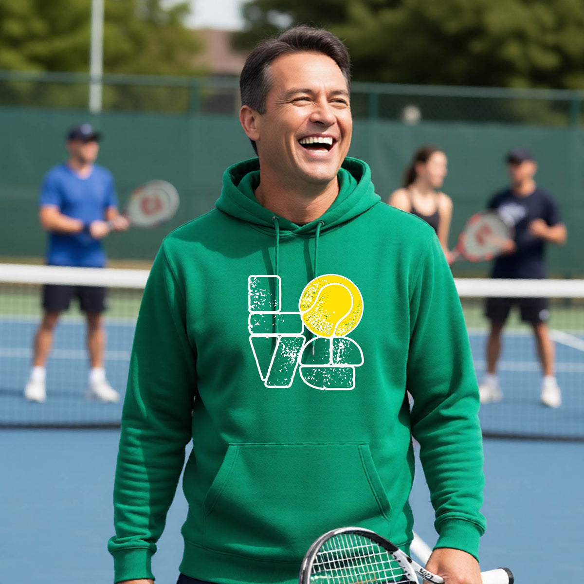 A smiling man wearing a green hoodie with a 'LOVE' tennis graphic with a tennis ball replacing the O, stands on an outdoor tennis court, with other players blurred in the background