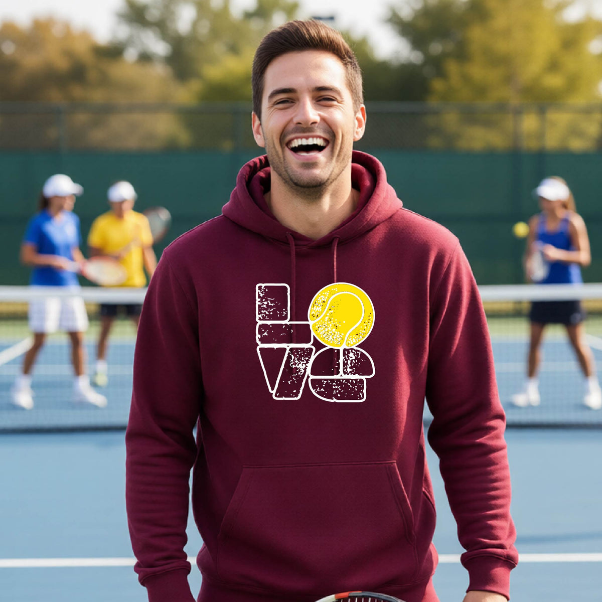 A smiling man wearing a maroon hoodie with a 'LOVE' tennis graphic with a tennis ball replacing the O, stands on an outdoor tennis court, with other players blurred in the background