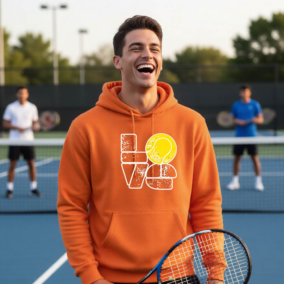 A smiling man wearing an orange hoodie with a 'LOVE' tennis graphic with a tennis ball replacing the O, stands on an outdoor tennis court, with other players blurred in the background