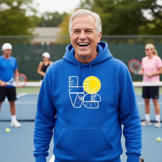 A smiling older man wearing a bright blue hoodie with a 'LOVE' tennis graphic with a tennis ball replacing the O, stands on an outdoor tennis court, with other players blurred in the background.