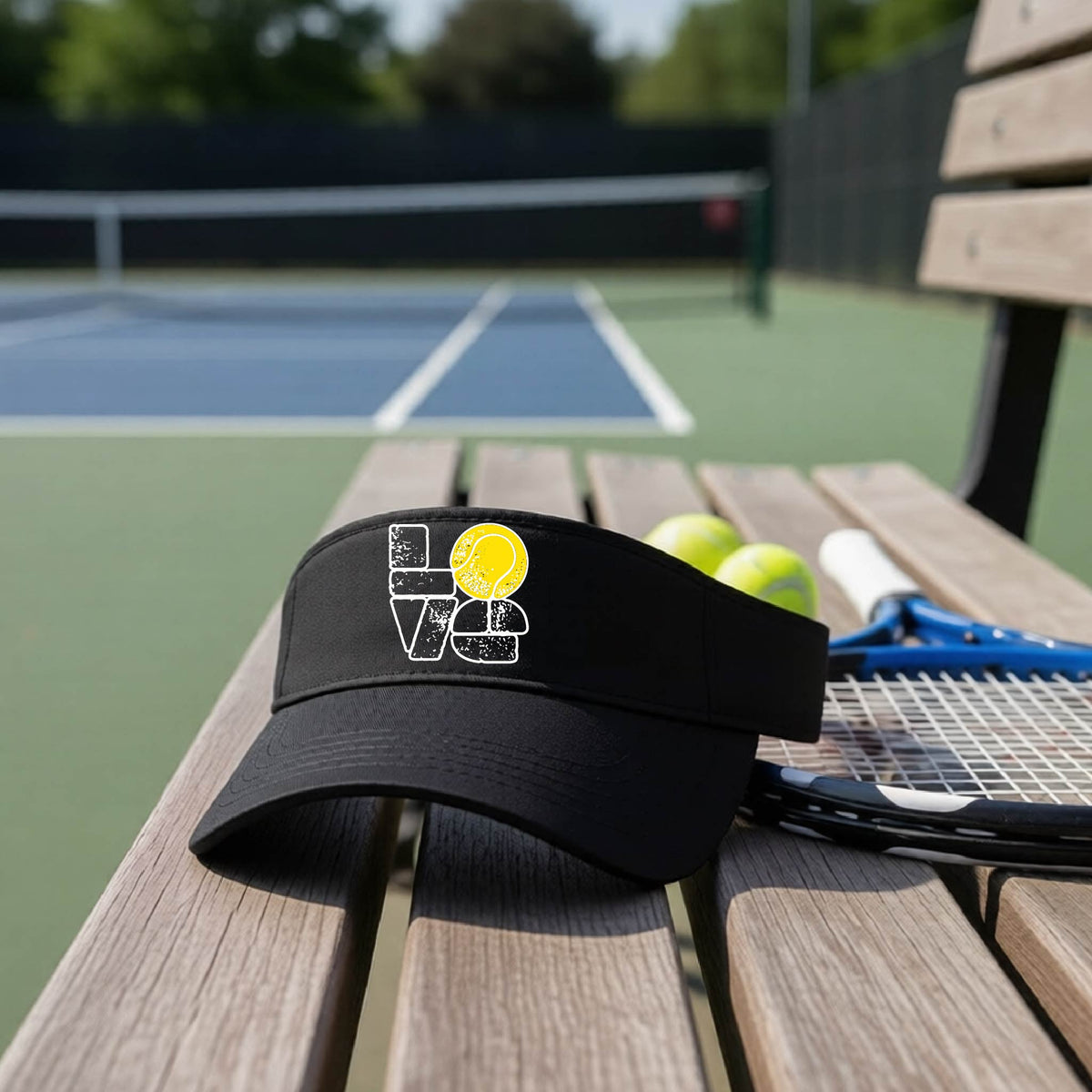 A black visor with a white and yellow tennis-themed graphic "LOVE" with a tennis ball replacing the O, sits on a wooden bench at a tennis court, with tennis balls and a racket in the background.
