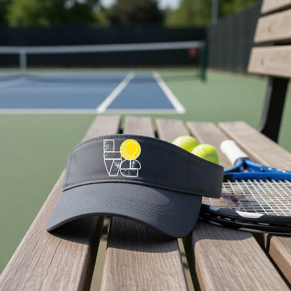 A graphite gray visor with a white and yellow tennis-themed graphic "LOVE" with a tennis ball replacing the O, sits on a wooden bench at a tennis court, with tennis balls and a racket in the background.