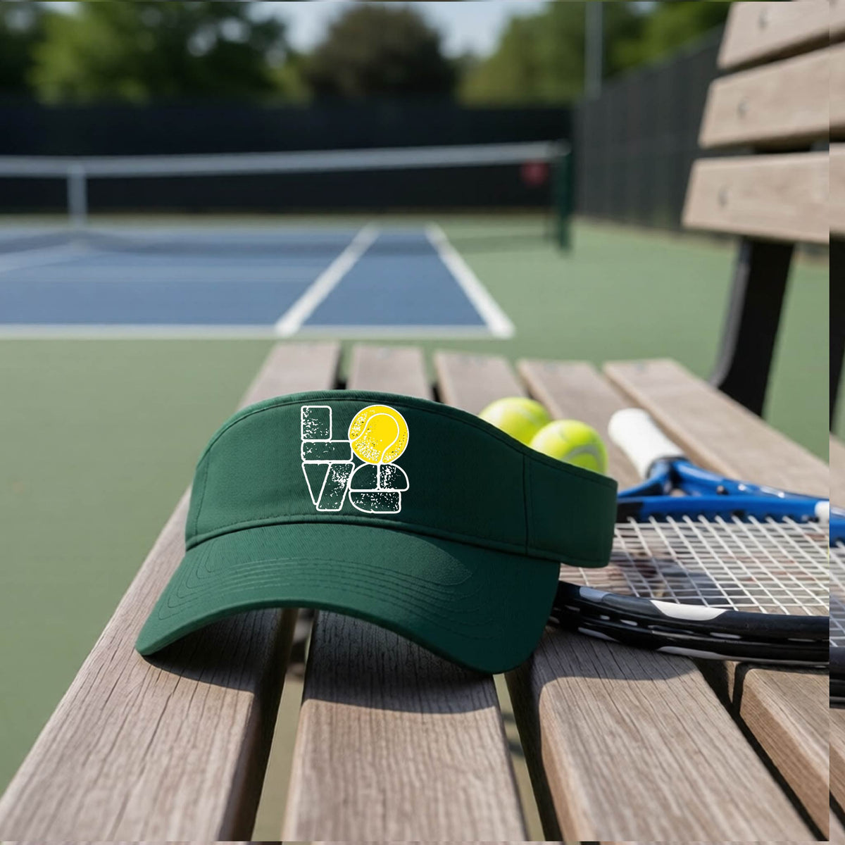 A green visor with a white and yellow tennis-themed graphic "LOVE" with a tennis ball replacing the O, sits on a wooden bench at a tennis court, with tennis balls and a racket in the background.